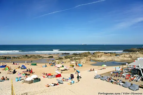 Praia de Leça da Palmeira com areal, banhistas ao longe, rochas e mar azul em dia de sol