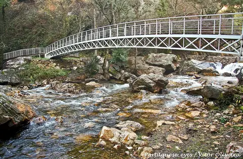 Ponte pedonal branca sobre ribeiro de águas límpidas e rochas nas Fragas de São Simão