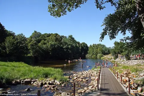 Praia fluvial da Aldeia Viçosa com passadiço de madeira, rio e banhistas ao longe entre árvores