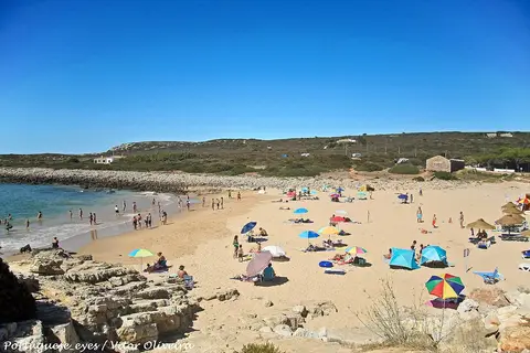 Praia da Ingrina com areal, chapéus de sol coloridos e mar calmo sob céu azul