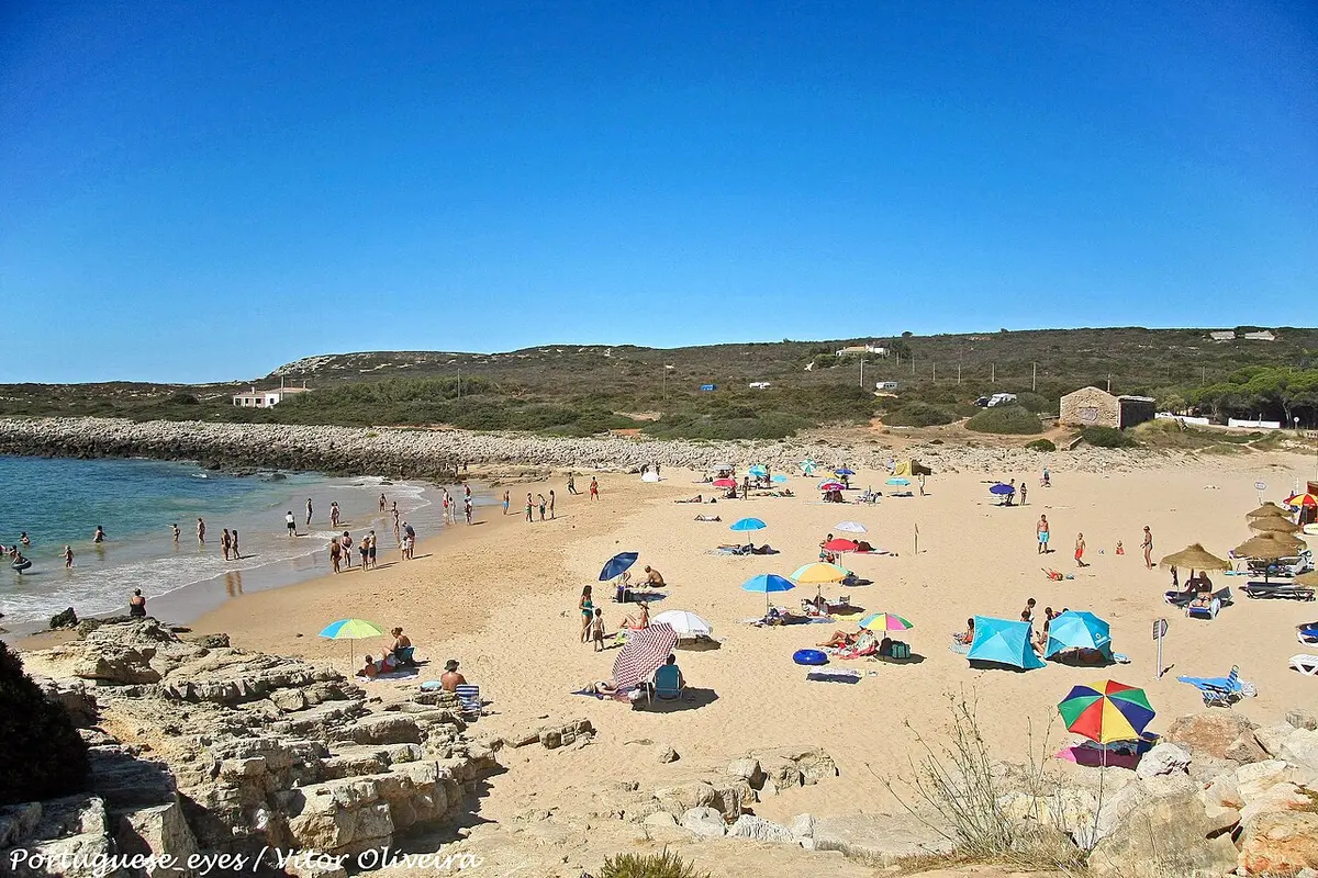 Praia da Ingrina com areal, chapéus de sol coloridos e mar calmo sob céu azul
