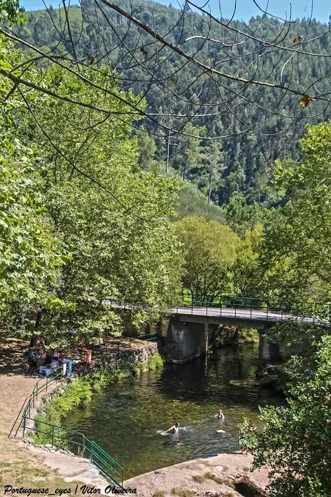 Praia fluvial com poço de água, ponte de betão e vegetação densa em redor