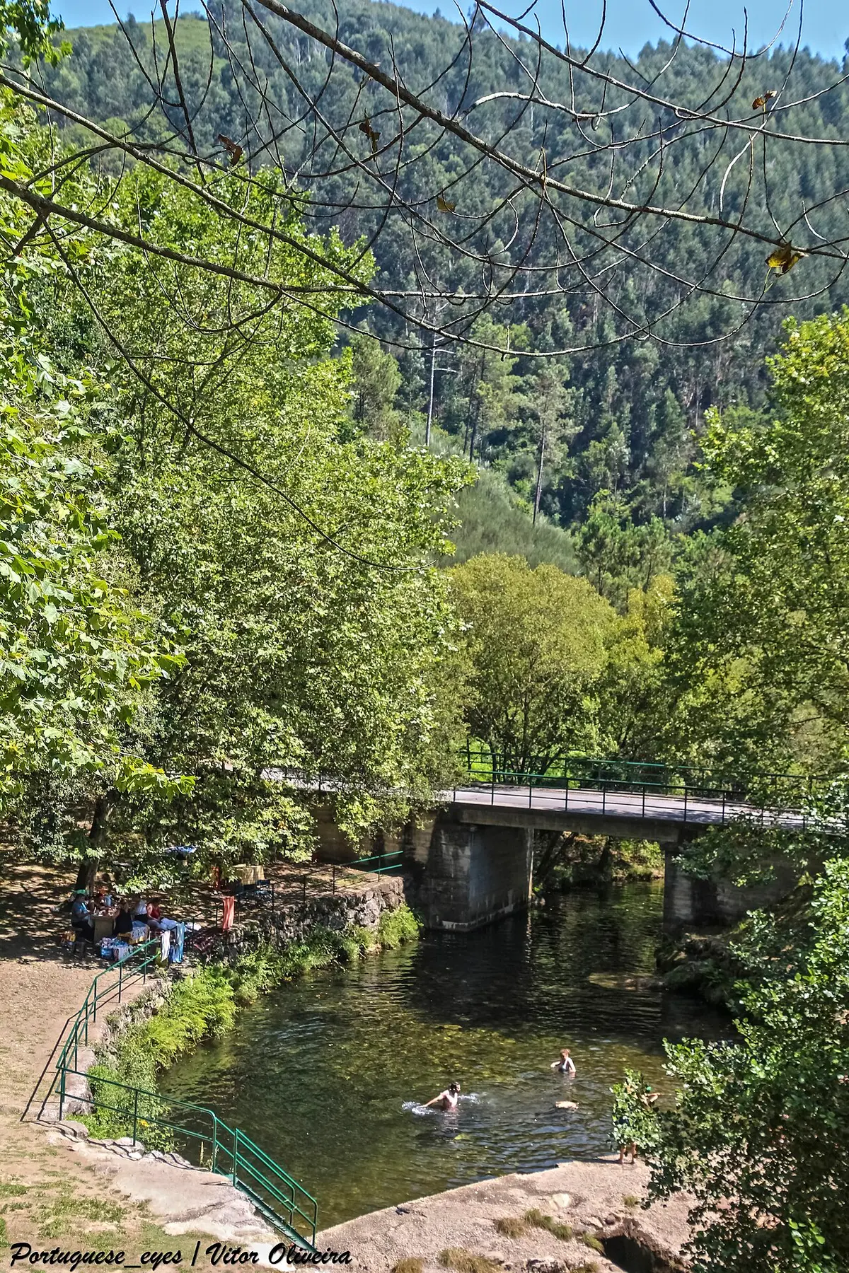 Praia fluvial com poço de água, ponte de betão e vegetação densa em redor