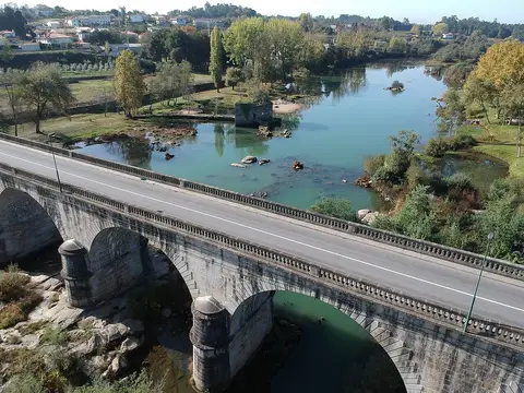 Vista aérea da Ponte do Bico, em pedra, sobre o rio com margens verdes e praia fluvial