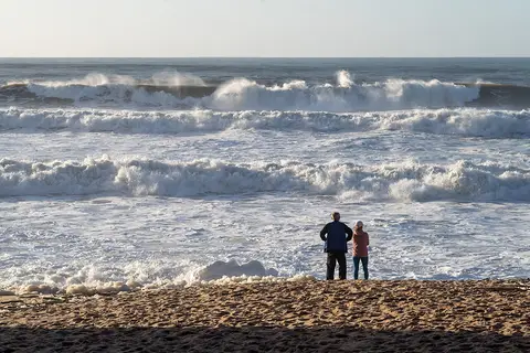 Duas pessoas de costas na areia da Praia de Valmitão a observar as ondas do mar agitado.