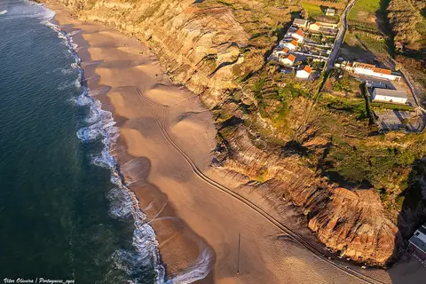 Vista aérea da Praia do Valmitão, com areal, ondas e falésias junto a casas no topo