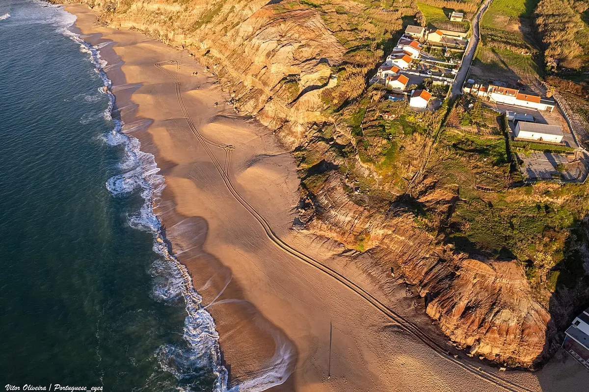 Vista aérea da Praia do Valmitão, com areal, ondas e falésias junto a casas no topo