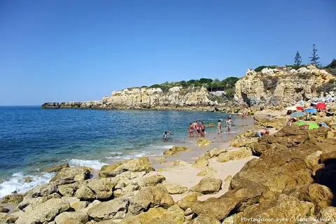 Praia do Castelo com rochas douradas, mar azul e banhistas ao longe junto à falésia