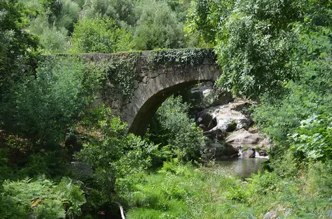 Ponte de pedra em arco sobre ribeiro rochoso, rodeada de vegetação, em Cavez