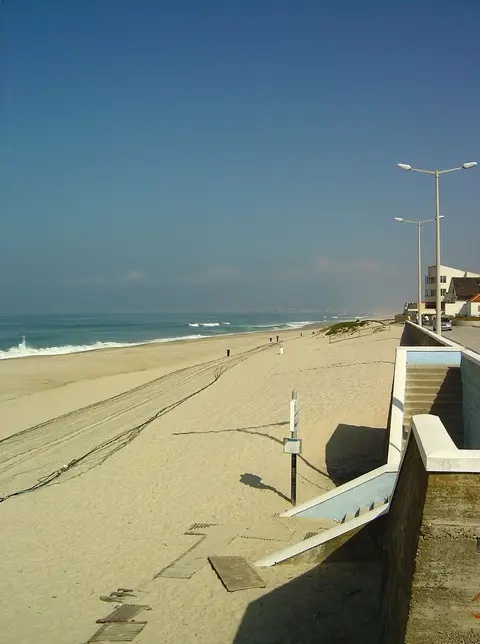 Praia extensa de areia com acesso em rampa e escadas junto ao passeio marítimo, mar calmo ao fundo