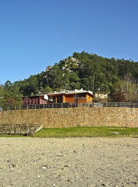 Praia fluvial do Reconquinho com muro de pedra, edifício de apoio em madeira e encosta arborizada