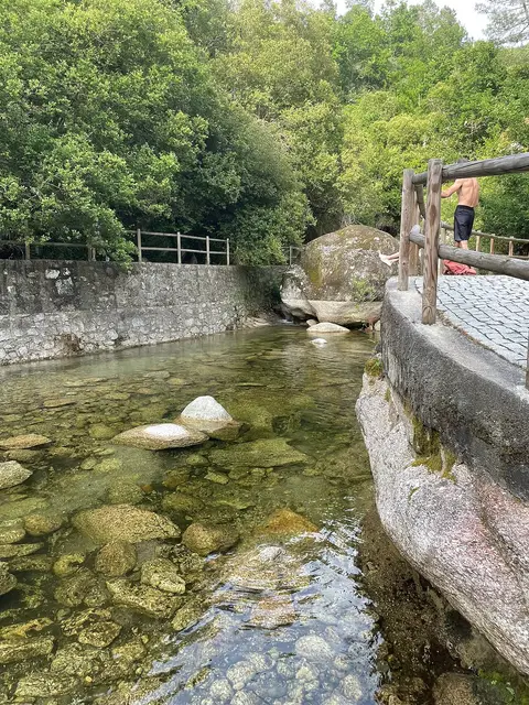 Praia fluvial de Lapa dos Dinheiros, com água transparente sobre pedras e muros de granito rodeados de vegetação