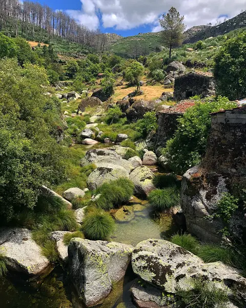 Ribeira de águas claras entre grandes rochas graníticas e vegetação, junto a casas de pedra na Serra da Estrela