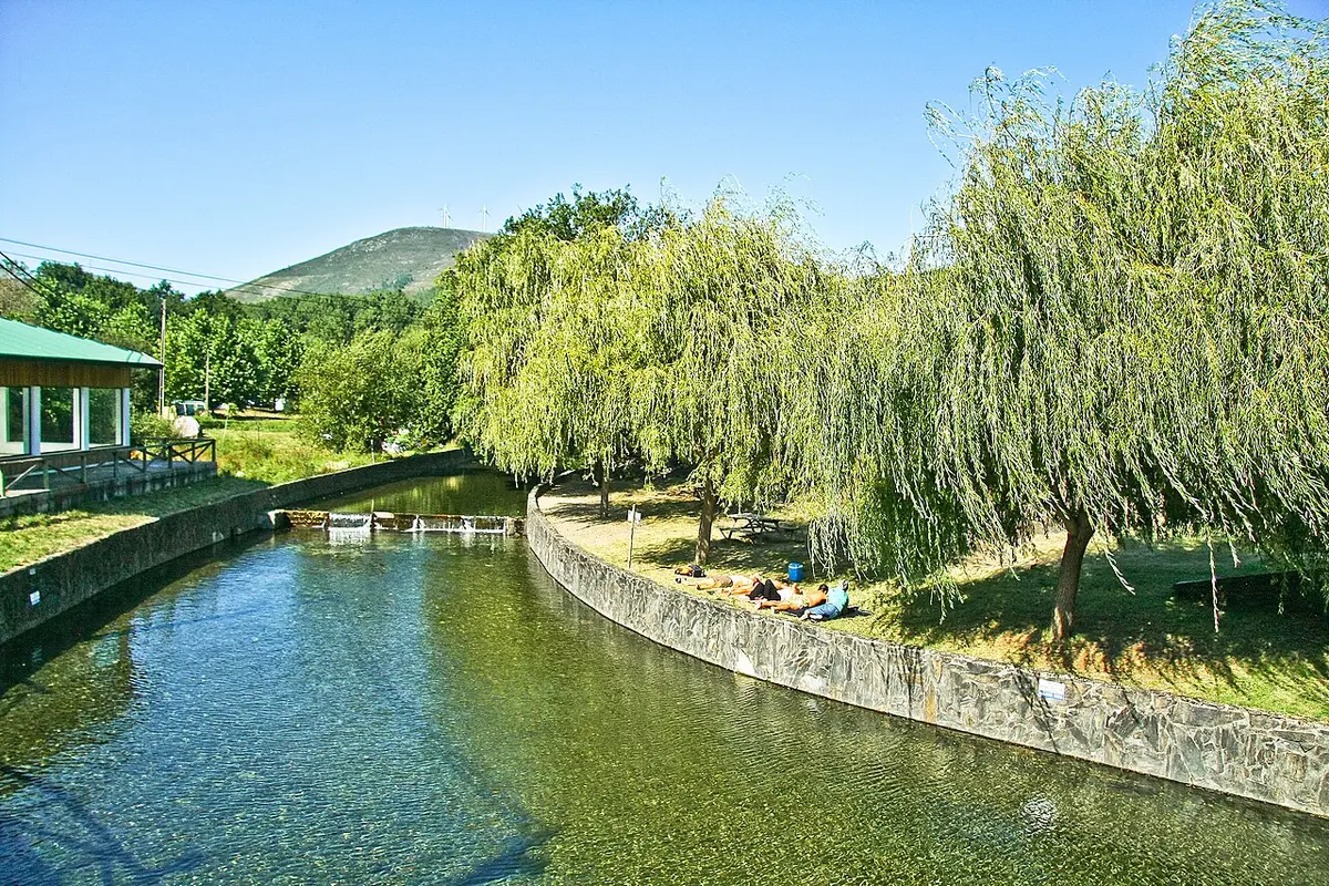 Praia fluvial do Poço da Corga, com rio de águas claras, salgueiros-chorões e relvado à beira-rio