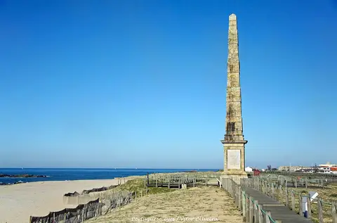 Padrão da Memória, obelisco de pedra junto à praia, com passadiço de madeira e mar ao fundo