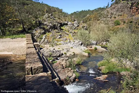 Praia fluvial do Penedo Furado com açude de pedra, rochas e vegetação junto ao rio