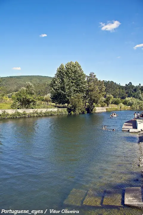 Praia fluvial da Senhora da Graça em Serpins, com rio, banhistas ao longe e vegetação na margem