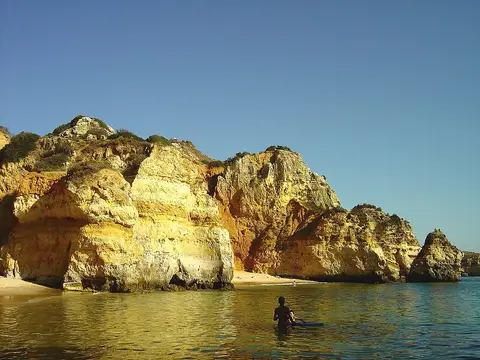 Falésias douradas e mar calmo na Praia do Camilo, com uma pessoa de costas na água