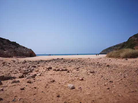Praia das Furnas com areia e pedras em primeiro plano, falésias e mar azul ao fundo