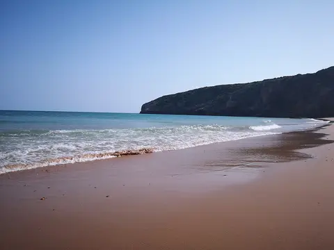 Praia das Furnas com areia dourada, ondas suaves e falésia verde ao fundo sob céu limpo