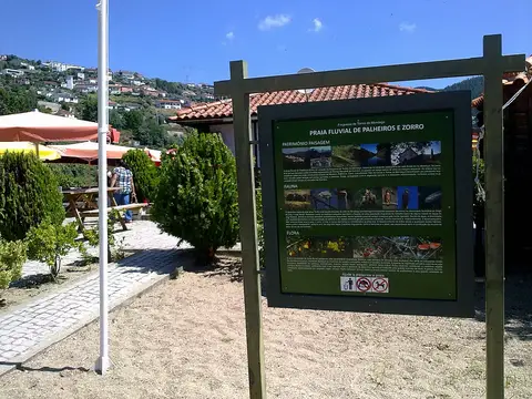 Painel informativo da Praia Fluvial de Palheiros e Zorro, com esplanada e casas ao fundo