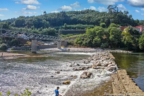 Praia fluvial com açude de pedras no rio, ponte metálica e encosta arborizada ao fundo
