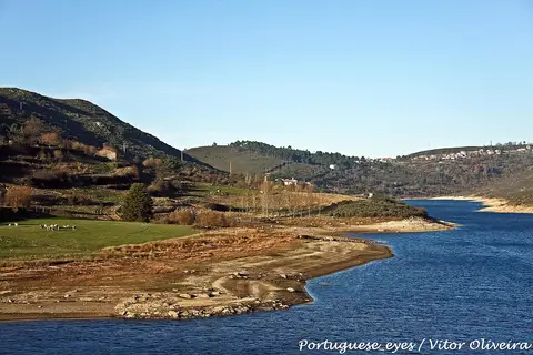 Albufeira do Caldeirão com margens de areia, colinas verdes e aldeia ao fundo sob céu azul