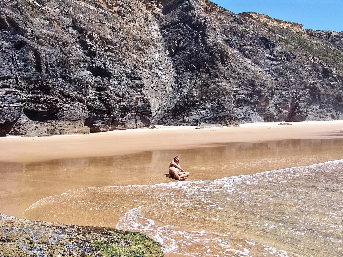 Praia de areia junto a falésias escarpadas, com água a chegar à areia e uma pessoa sentada ao longe