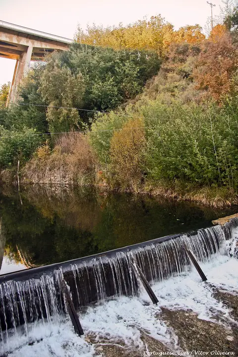 Açude com queda de água na praia fluvial de Mega Fundeira, rodeado de vegetação e ponte ao fundo