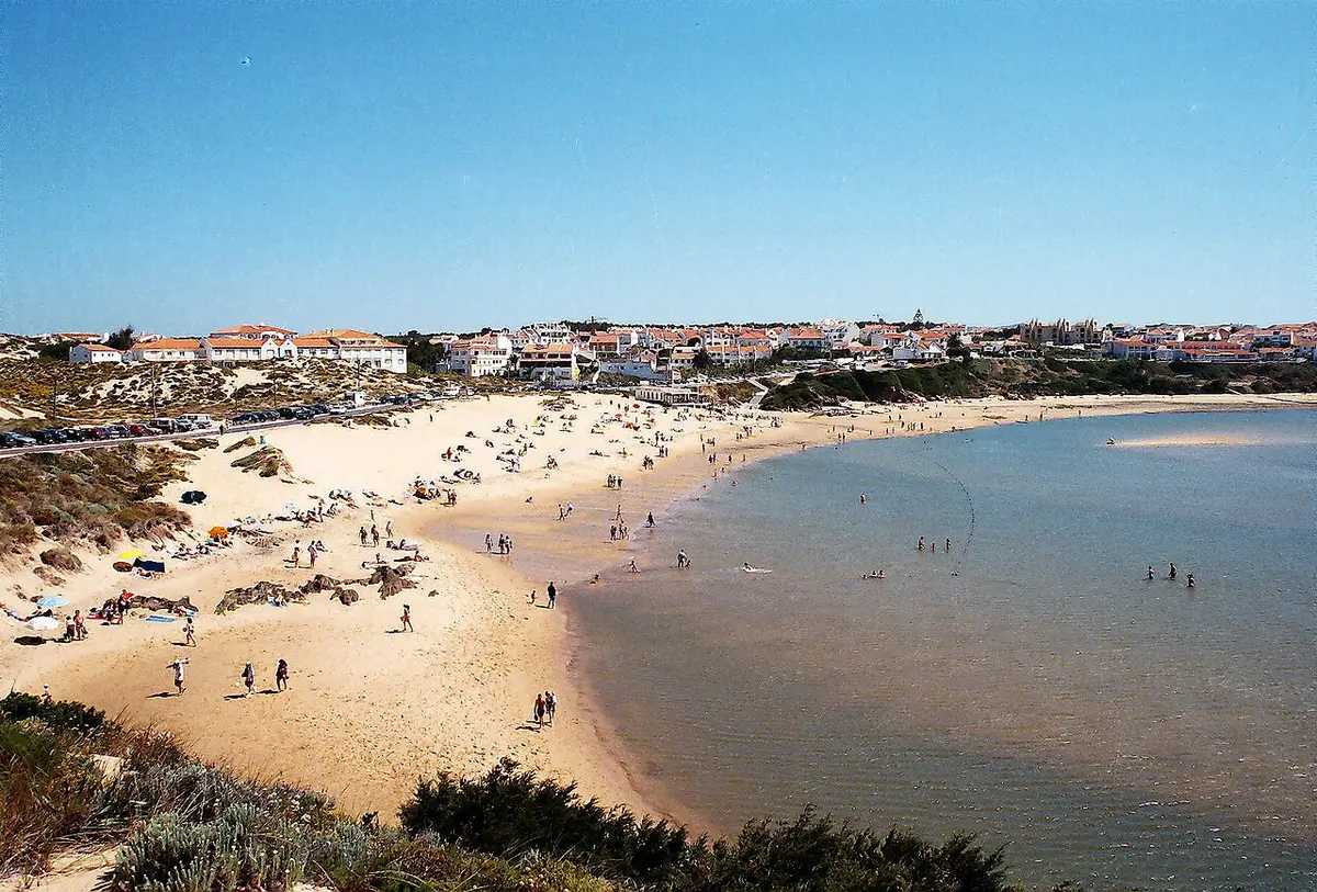 Praia da Franquia em Vila Nova de Milfontes, com areal, banhistas ao longe e a vila ao fundo