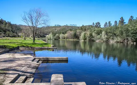 Praia fluvial de Cambas com plataformas de cimento junto à água calma e encostas arborizadas ao fundo
