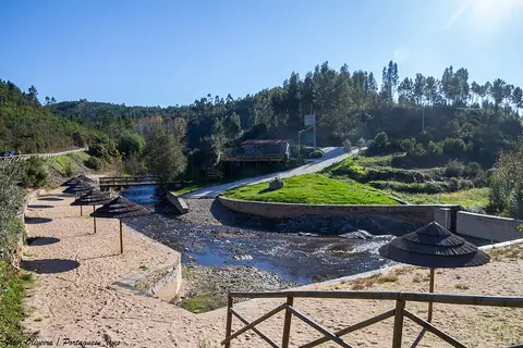 Praia fluvial do Boiçô com areal, guarda-sóis de colmo e ribeiro rodeado de colinas arborizadas