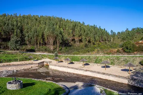Praia fluvial do Boiçô com guarda-sóis de colmo na margem arenosa, junto a encosta de eucaliptos.
