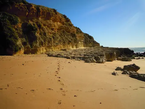 Praia dos Aveiros com areia dourada, pegadas e falésias rochosas junto ao mar