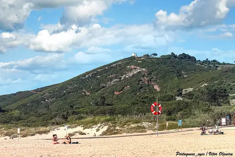 Praia de areia com boia salva-vidas e colina verde ao fundo, sob céu com nuvens
