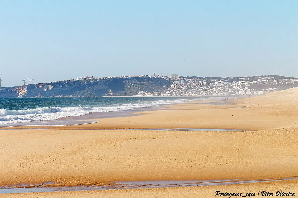 Praia do Salgado com areal extenso, ondas e a vila da Nazaré ao fundo sobre a falésia
