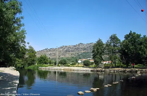 Praia fluvial no rio Mondego com margens de pedra, árvores e serra ao fundo sob céu azul