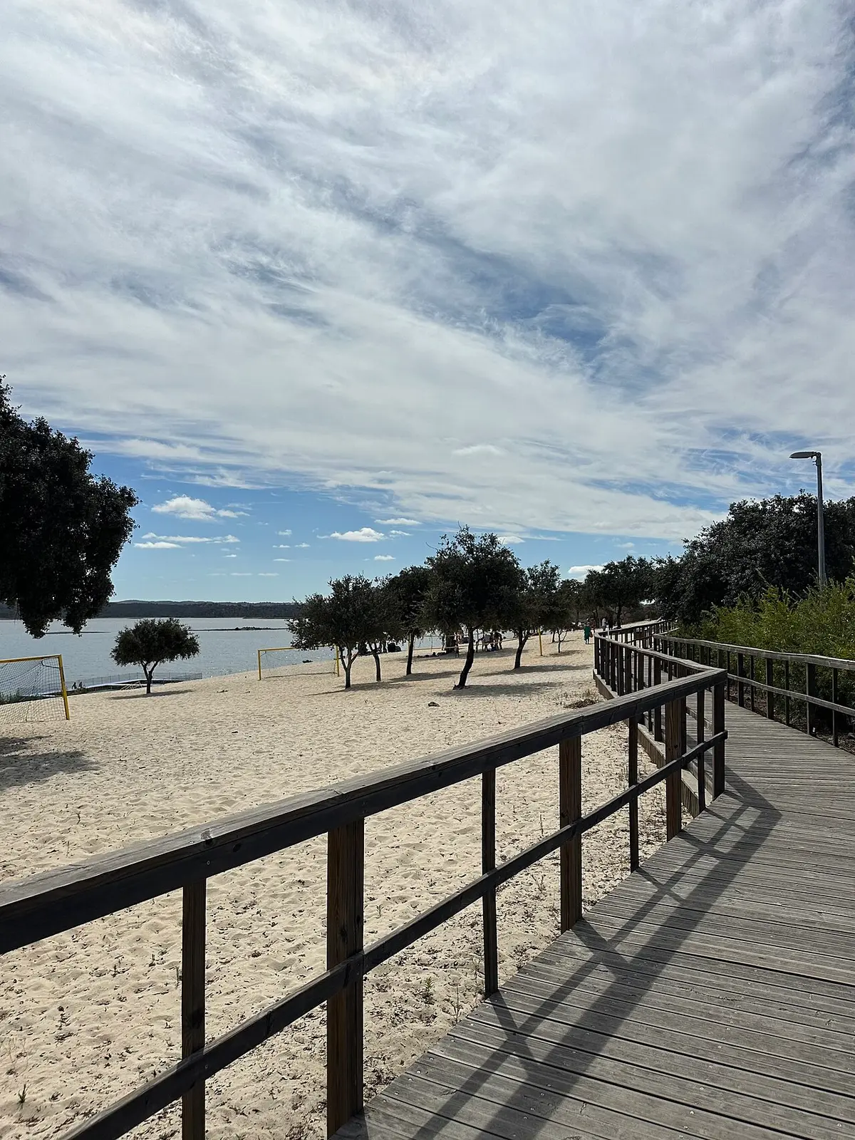 Passadiço de madeira junto à praia fluvial de areia, com árvores e albufeira ao fundo