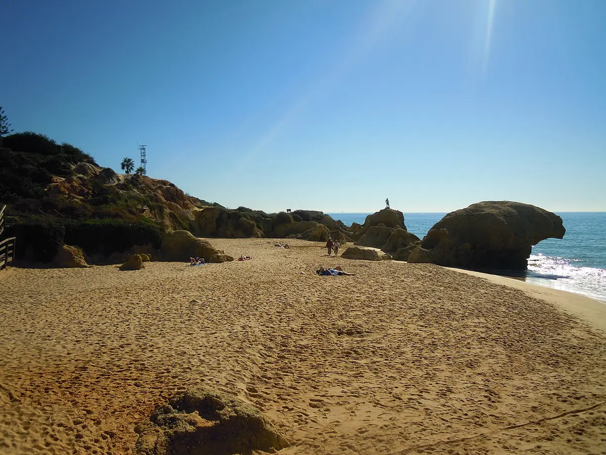 Praia da Galé com areal amplo, formações rochosas douradas e mar calmo sob céu azul