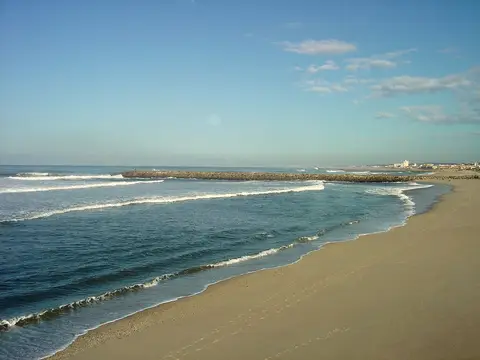 Praia de areia em Paramos com ondas suaves e quebra-mar de pedra ao fundo, sob céu azul