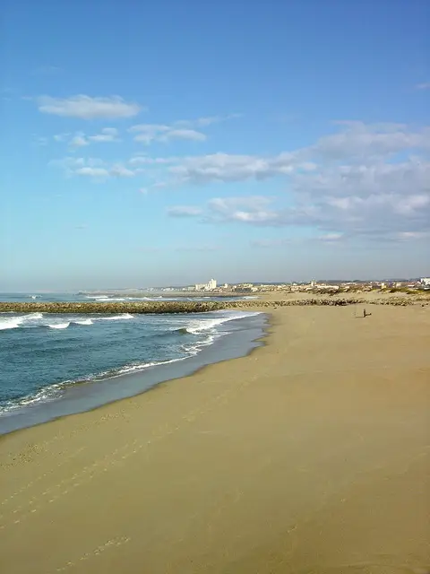 Praia de areia em Paramos com quebra-mar, ondas e a povoação ao fundo sob céu azul