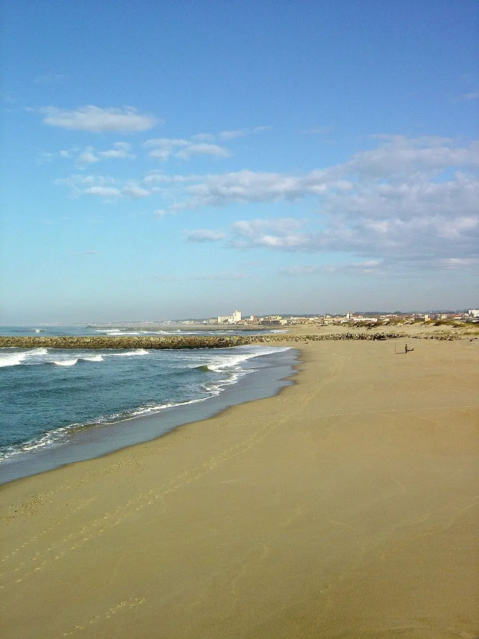 Praia de areia em Paramos com quebra-mar, ondas e a povoação ao fundo sob céu azul