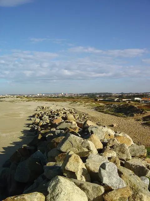Praia de Paramos com enrocamento de pedras sobre a areia e dunas, sob céu azul com nuvens