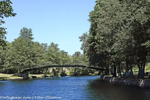 Rio calmo com ponte pedonal metálica e margens arborizadas na praia fluvial de Segões