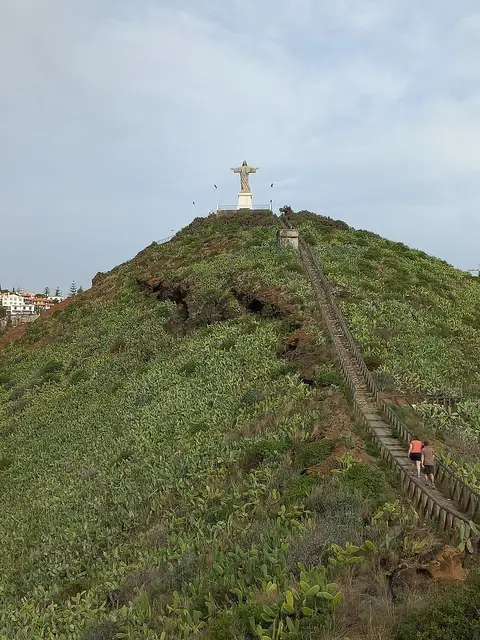Estátua do Cristo Rei no topo da colina verde da Ponta do Garajau, com escadaria a subir a encosta