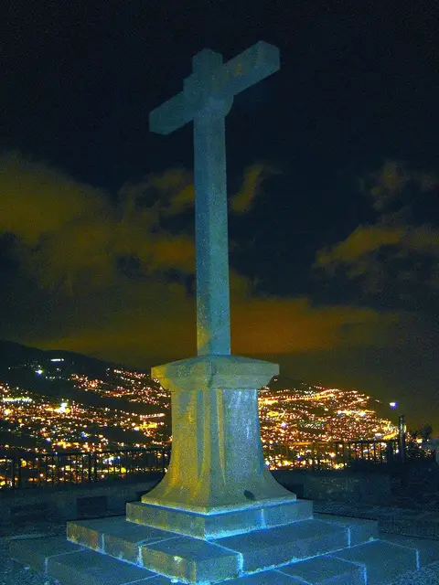 Cruz de pedra no miradouro do Pico dos Barcelos à noite, com as luzes do Funchal ao fundo