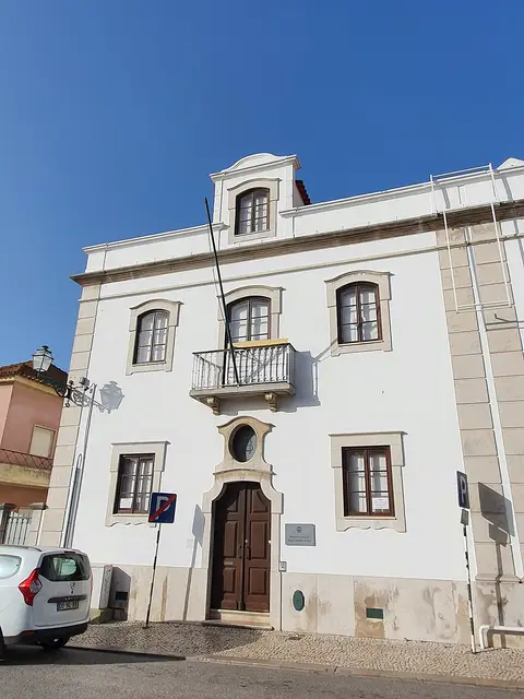 Fachada branca de edifício histórico de dois pisos com varanda e porta de madeira, sede da biblioteca municipal
