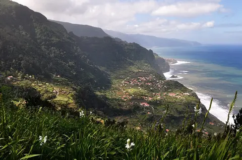 Vista panorâmica de miradouro sobre encostas verdes, aldeia costeira e oceano na Madeira