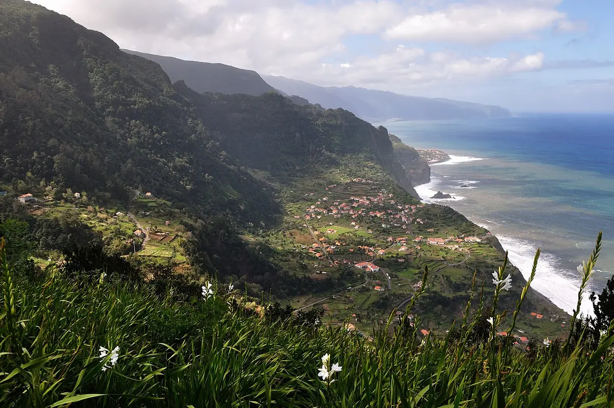 Vista panorâmica de miradouro sobre encostas verdes, aldeia costeira e oceano na Madeira