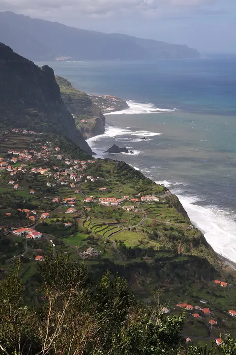 Vista panorâmica de miradouro sobre a costa norte da Madeira, com aldeia, terraços verdes e falésias sobre o mar